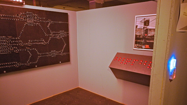 A room corner with a complex network of lights on the wall and a control panel with numerous red buttons