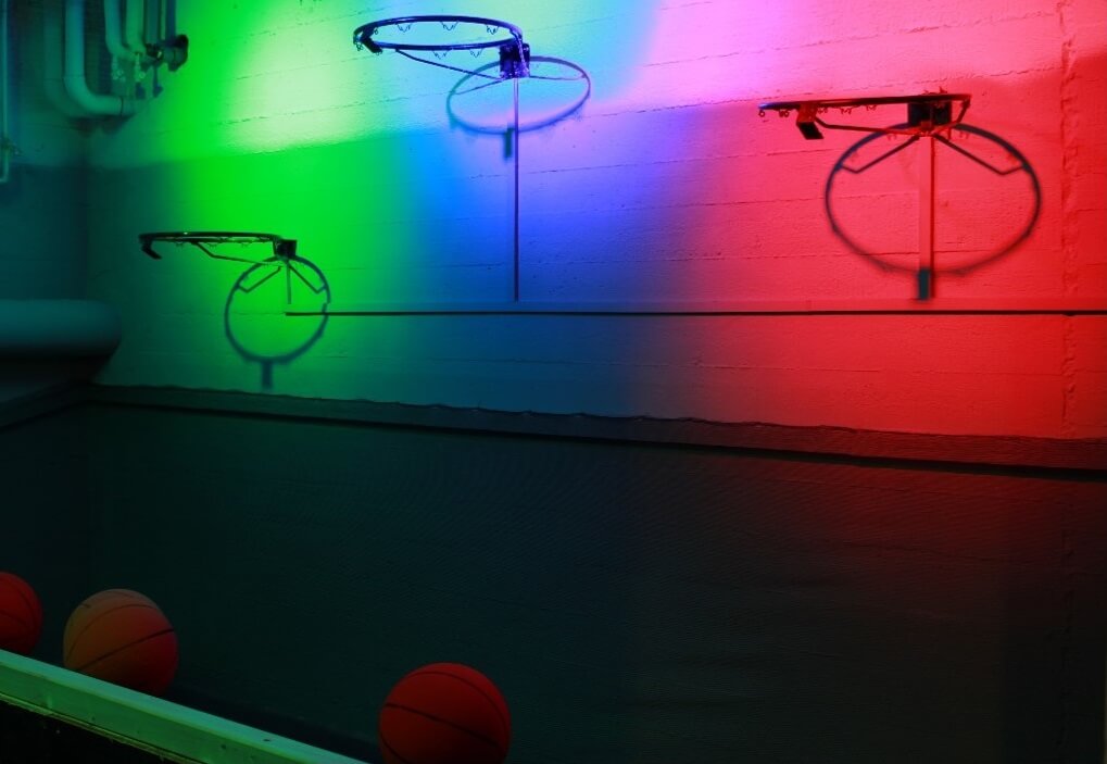 Three basketball hoops illuminated in different colours, with some basketballs underneath