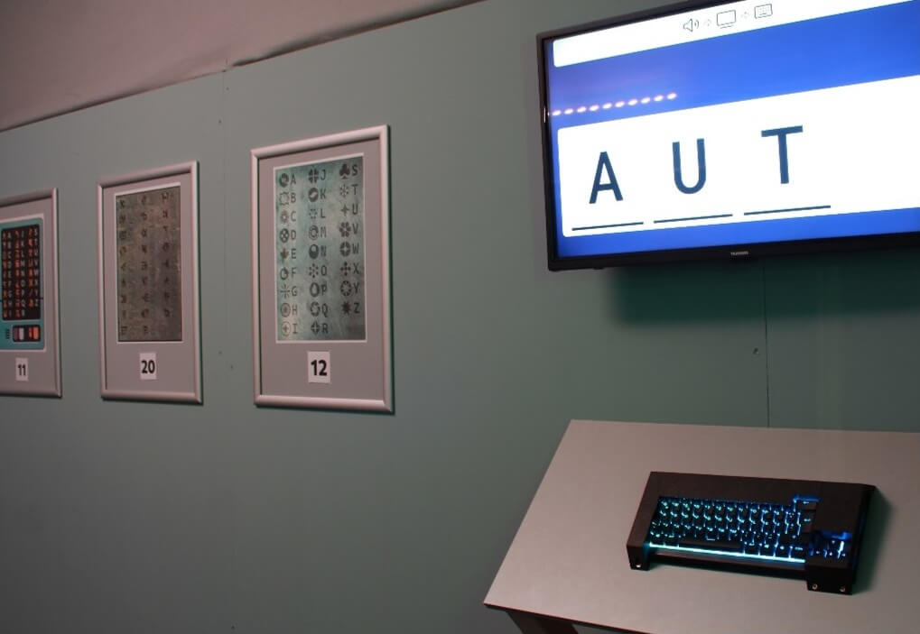 View into a room with posters featuring code tables on the walls, with a keyboard and a screen displaying the letters A, U, and T in the foreground