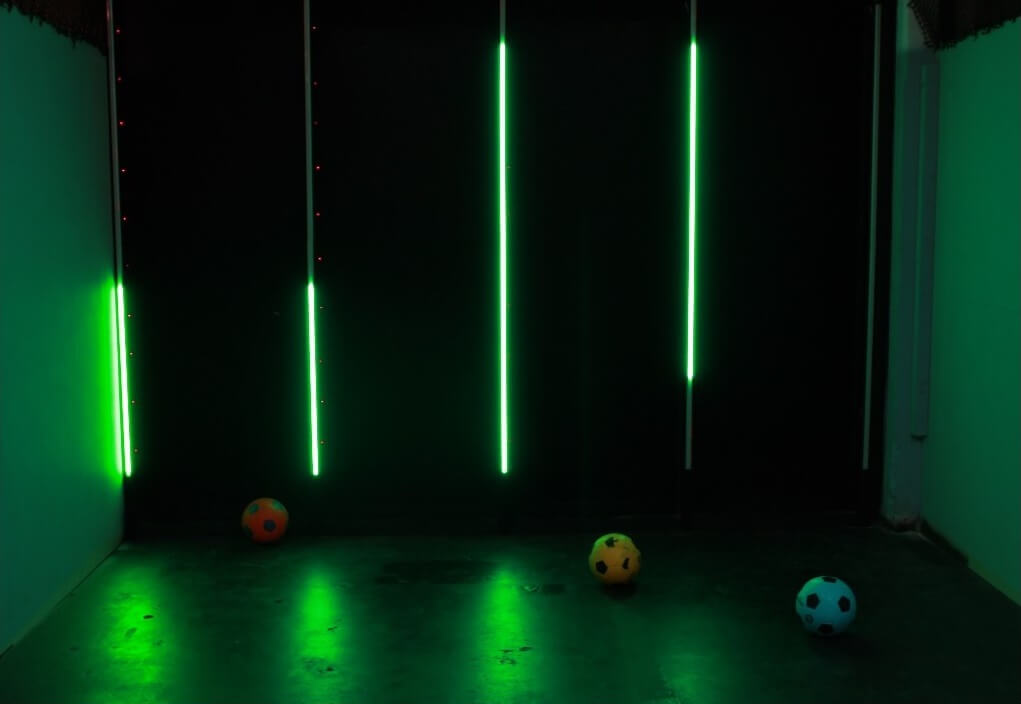 A wall with green vertical LED strips at varying heights from floor to ceiling, with a few footballs in front