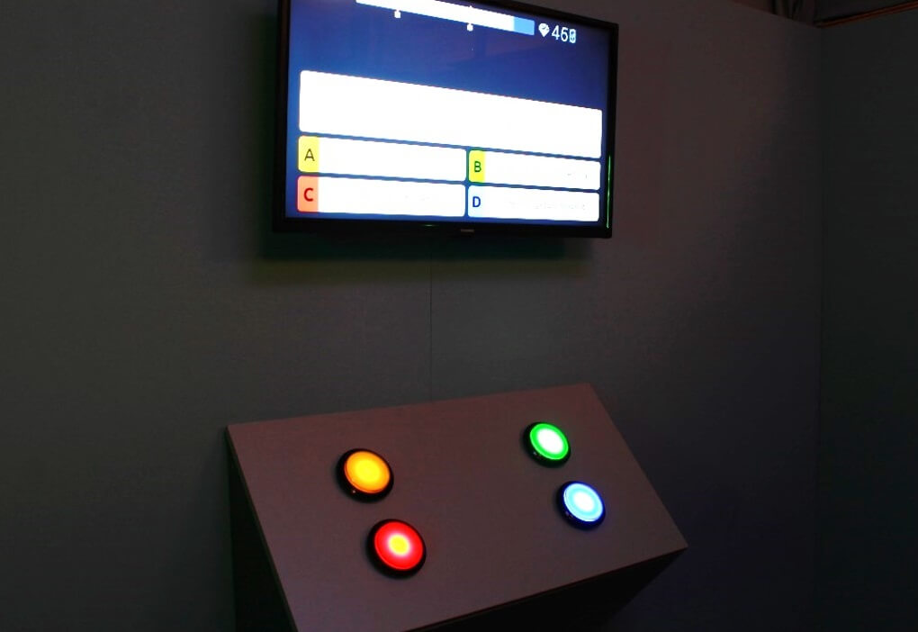A screen displaying a question with four answer options, below which is a console with four brightly glowing large round buttons