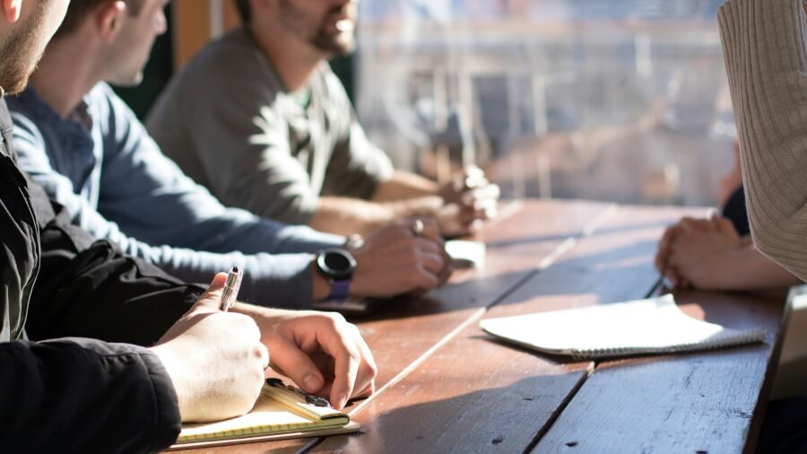 A close-up of a meeting table with hands holding pens and notebooks, indicating a collaborative discussion among participants